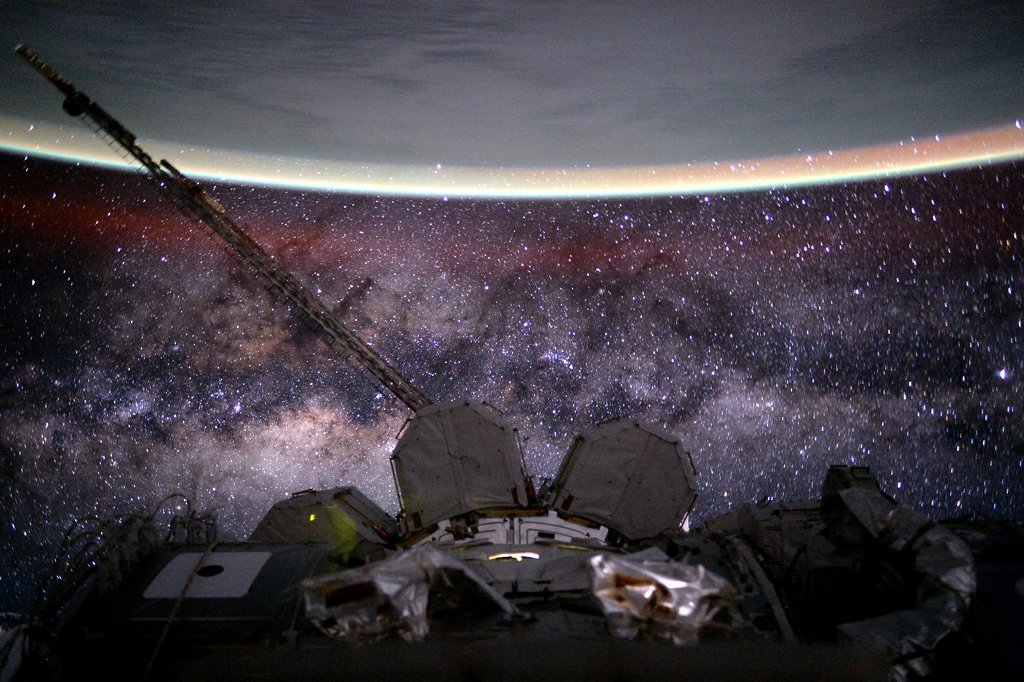 Über der Internationalen Raumstation breitet sich die Milchstraße mit hellen Sternfeldern und Dunkelwolken aus, von oben ragt die Erde ins Bild.