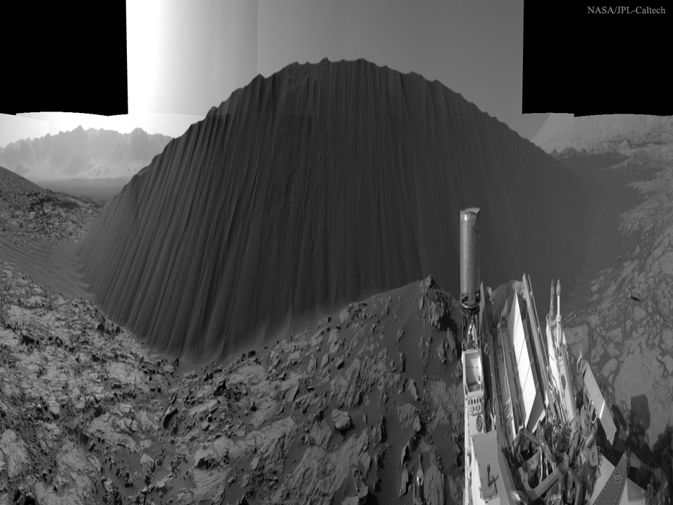 Das waagrecht komprimierte Bild zeigt eine dunkle Sanddüne, die im Krater Gale vor dem Rover Curiosity auf dem Mars aufragt. Links neben der Düne liegt Sand, der vom Wind verweht wurde. Das Gelände ist risig, helle Steine ragen aus dunklem Material.
