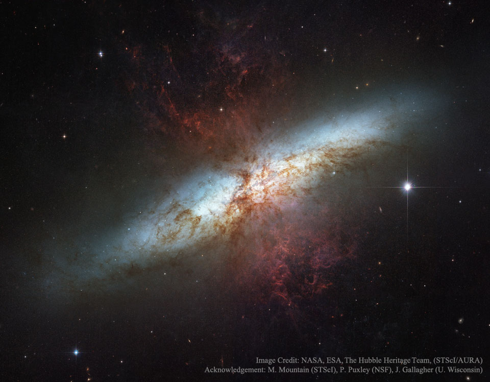 Die irreguläre Zigarrengalaxie M82 verläuft hell diagonal durchs Bild. In der Mitte leuchten dunkelrote Nebel, die an den Rauch einer Explosion erinnern.