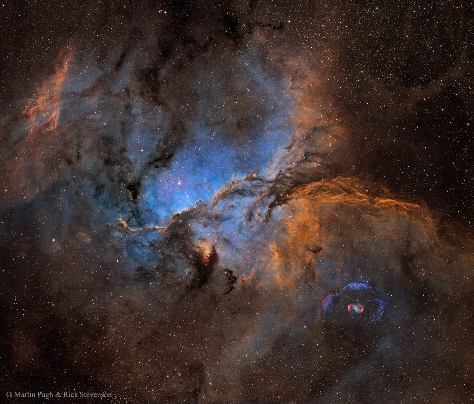 Mitten im Bild leuchtet ein blauer Hintergrund, über den dunkle Staubwolken verlaufen. das Blau ist von dunklen Nebeln umgeben, die teilweise in orange leuchtende Wolken übergehen.