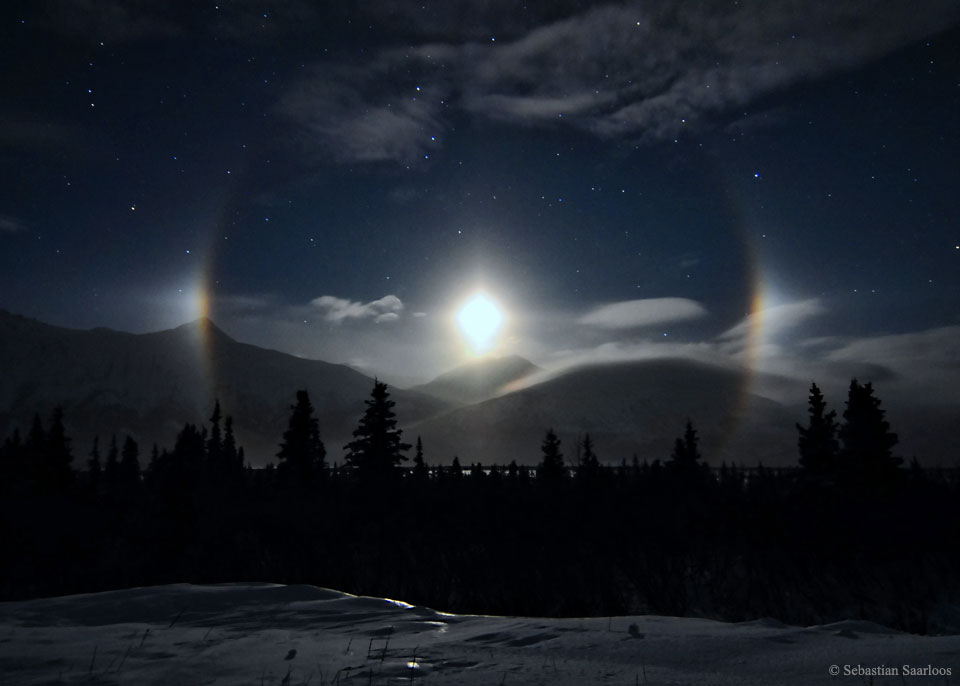 Über den Bergen am Lower Mill Creek in Alaska geht der Mond unter. Er ist von einem Hof umgeben, der links und rechts von Paraselena oder Nebenmonden flankiert sind.