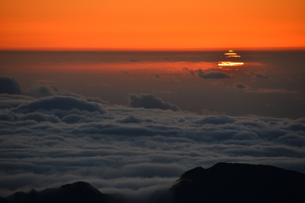 Hinter einer Wolkenbank geht die Sonne auf. Am oberen Rand leuchtet ein grüner Blitz. Der Aussichtspunkt ist die Caldera auf dem Haleakala auf der Insel Maui.