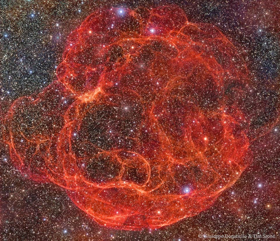 In einem dicht besiedelten Sternenfeld mit wenigen hellen Sternen leuchtet ein verworrenes Knäuel aus roten Strähnen, die von dunkleren roten Nebeln umgeben ist.