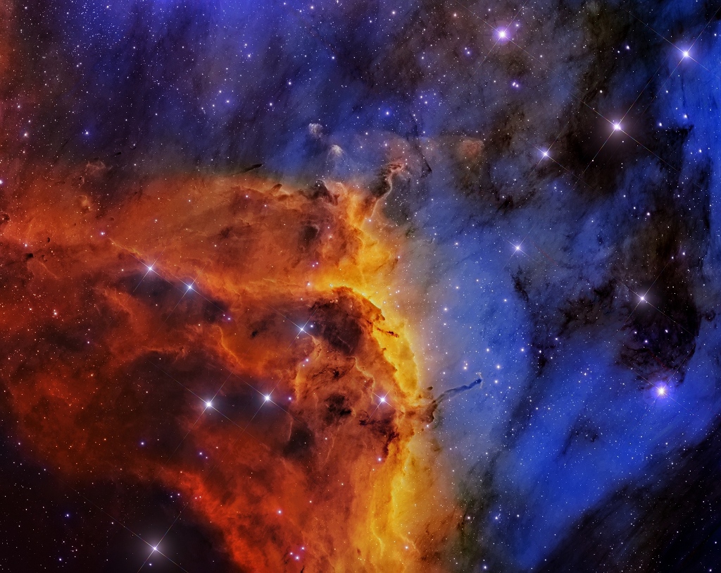 Von links unten ragt eine orange-braune aufgetürmte Wolke ins Bild, der Hintergrund ist schwarz-blau. Im Bild sind wenige Sterne verteilt.