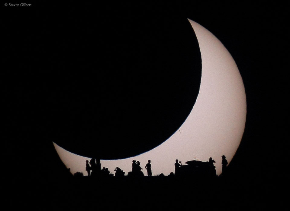 Der Mond bedeckt die Sonne, während sie aufgeht. Am Horizont sind die Silhouetten von Beobachtenden vor der Sonne zu sehen.