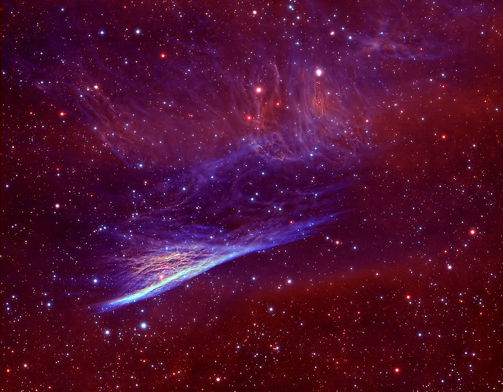 Vor einem rötlichen Nebel mit einem Teppich aus Sternen leuchtet ein blauer, strichförmiger Nebel, von dem nach oben Fasern auslaufen. Auch im Hintergrund sind einige Fasern erkennbar.