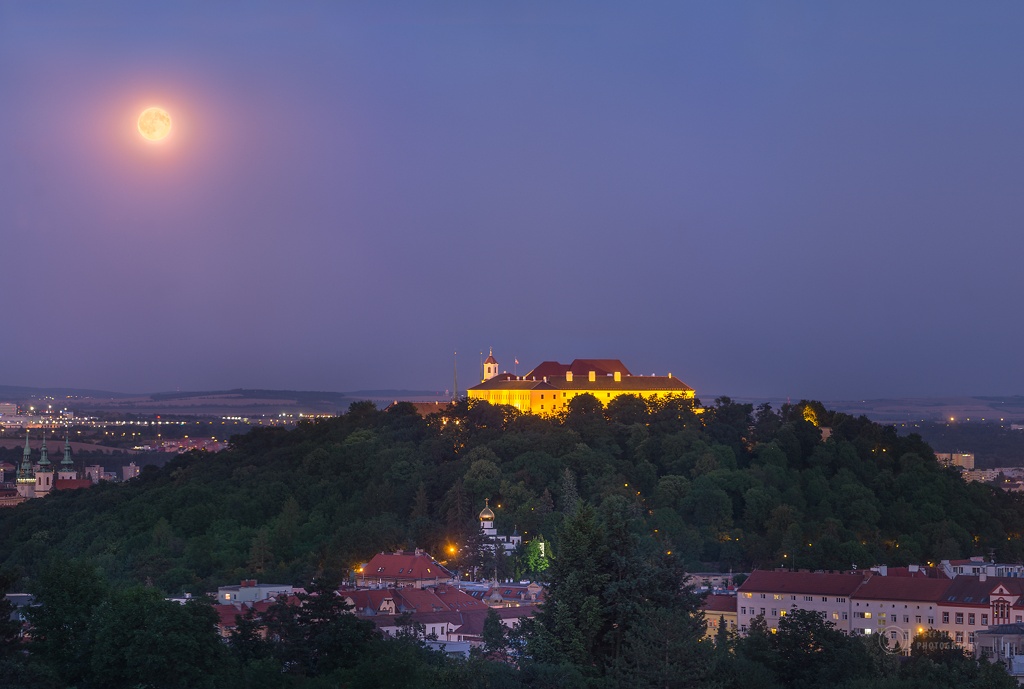 Auf einem bewaldeten Berg steht die beleuchtete Festung Špilberk über der tschechischen Stadt Brünn. LInkds darüber leuchtet der Vollmond.