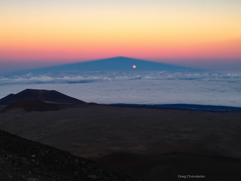 Hinter einem dunklen Gelände breitet sich eine Nebeldecke bis zum Horizont aus, dahinter steigt am Himmel im Abendrot der Schatten eines Vulkans auf. Im Schattenkegel leuchtet der aufgehende Mond.