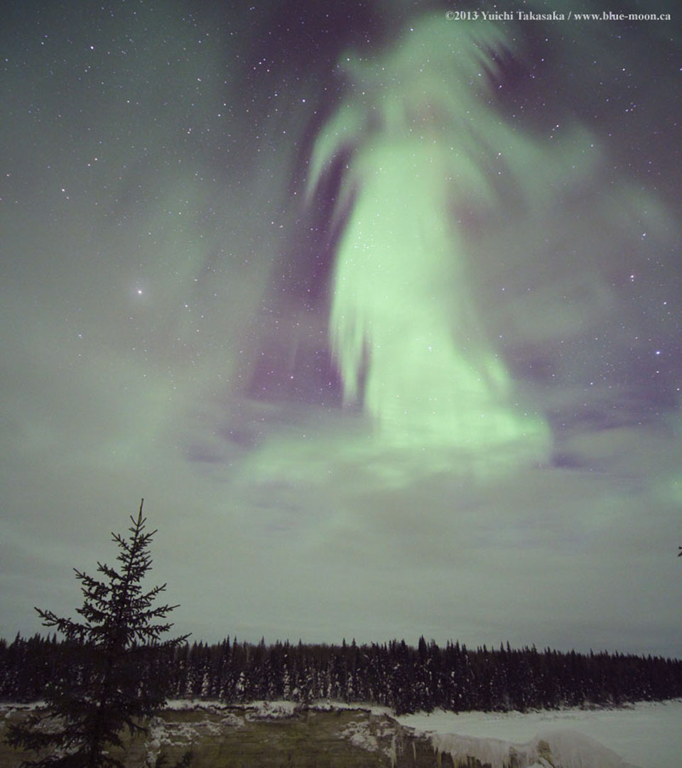 Über einer verschneiten Winterlandschaft steht am Horizont ein Wald, im Vordergrund ist ein gefrorener Wasserfall. Am Himmel leuchtet ein Polarlicht in Form einer Gestalt, die an eine Hexe oder eine Göttin erinnert.