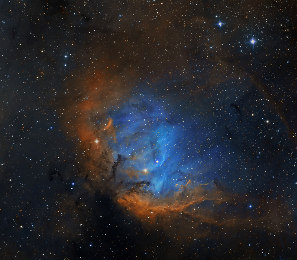 Ein orangefarbener Nebel ist schalenförmig nach rechts geöffnet, darin schimmert ein blauer, gefaserter Nebel, links unten sind zwei kleine dunkle Staubranken. Im Hintergrund sind viele kleine Sterne gleichmäßig verteilt.