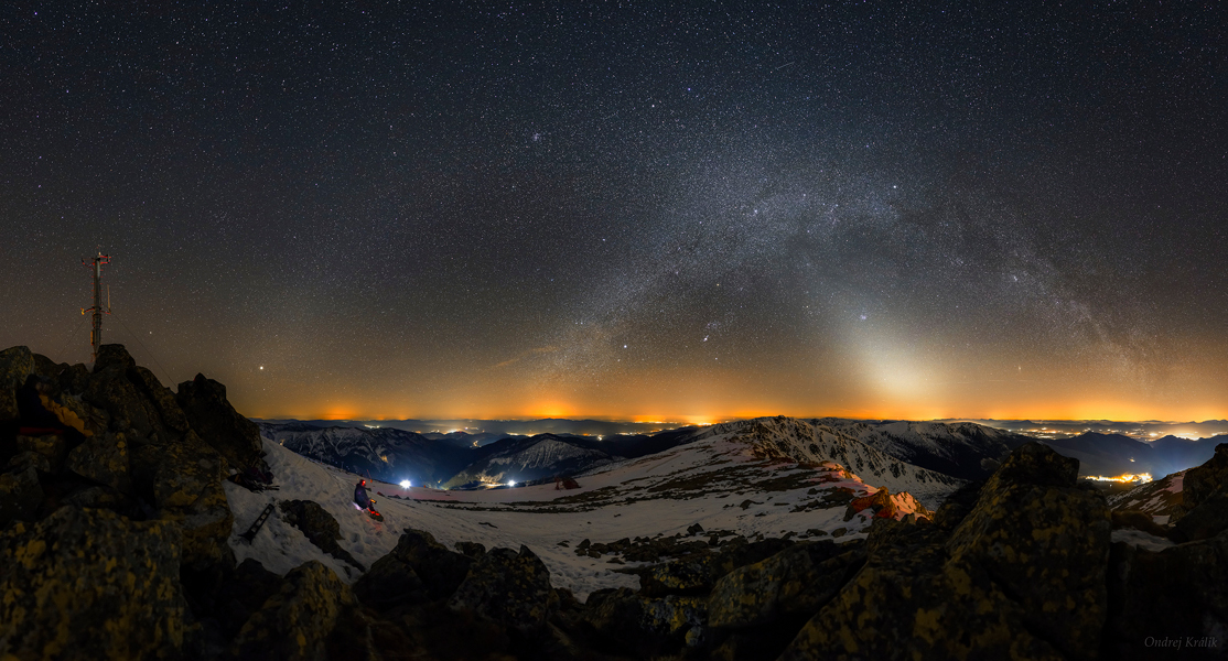 Über einem verschneiten Bergpanorama wölbt sich rechts die Milchstraße, der linke Bogen ist das schwache Band des Zodiakallichtes. Am Horizont leuchten helle orangefarbene Lichter.