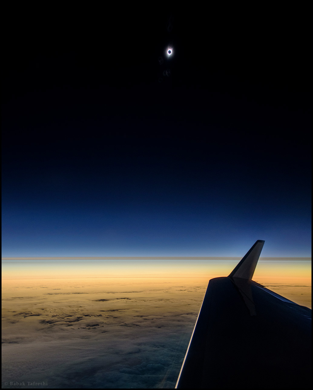 Hinter dem Flügel eines Flugzeugs sind Wolken, vorne im Schatten, hinten orangefarben. Am glasklaren, dunklen Himmel strahlt die Korona bei einer Sonnenfinsternis um den Neumond.