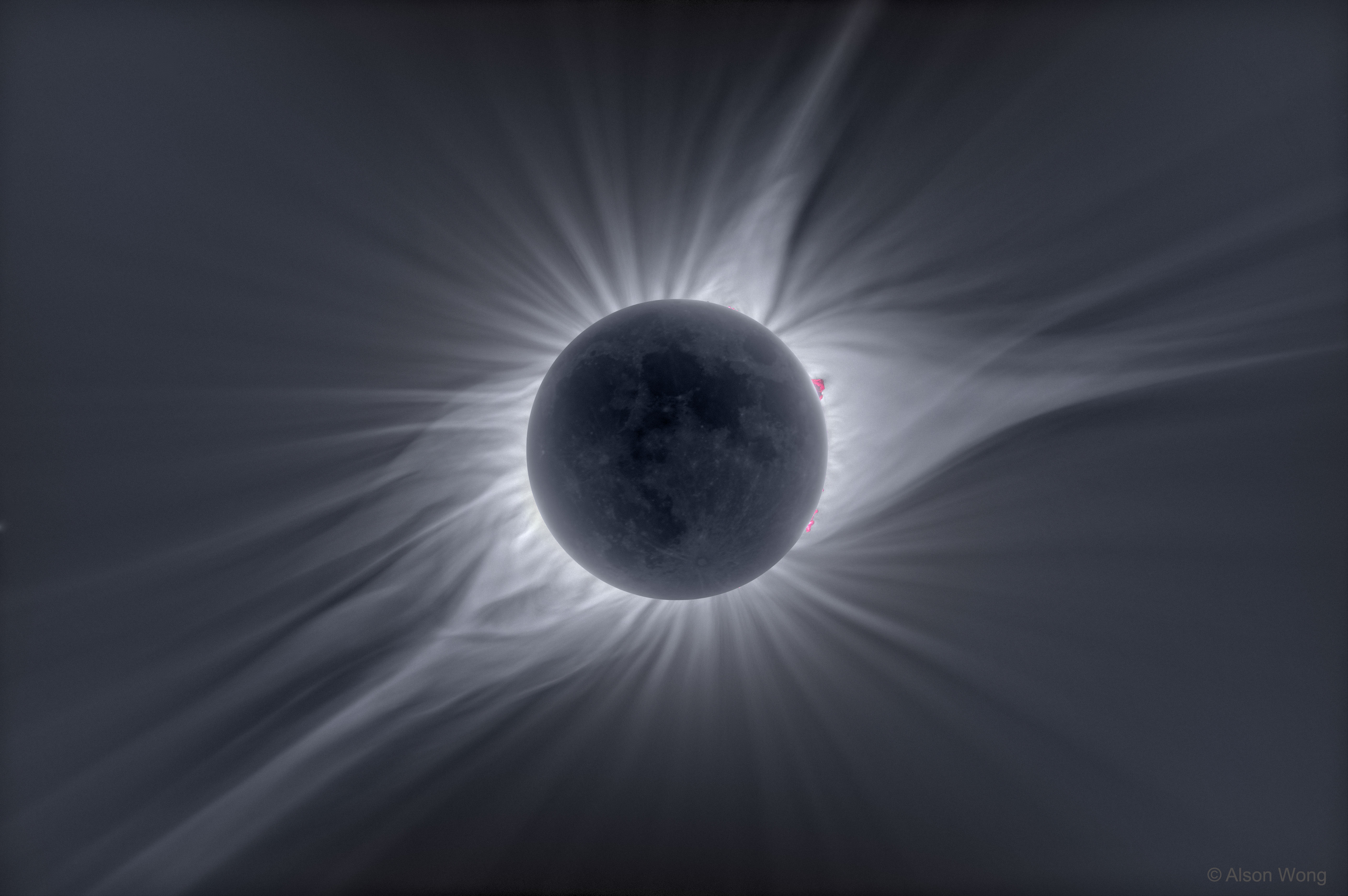 Mitten im Bild ist der schwach beleuchtete Mond bei einer totalen Sonnenfinsternis von den breit gefächerten Strahlen der Sonnenkorona umgeben. Am Rand ragen einige rosarote Protuberanzen auf.