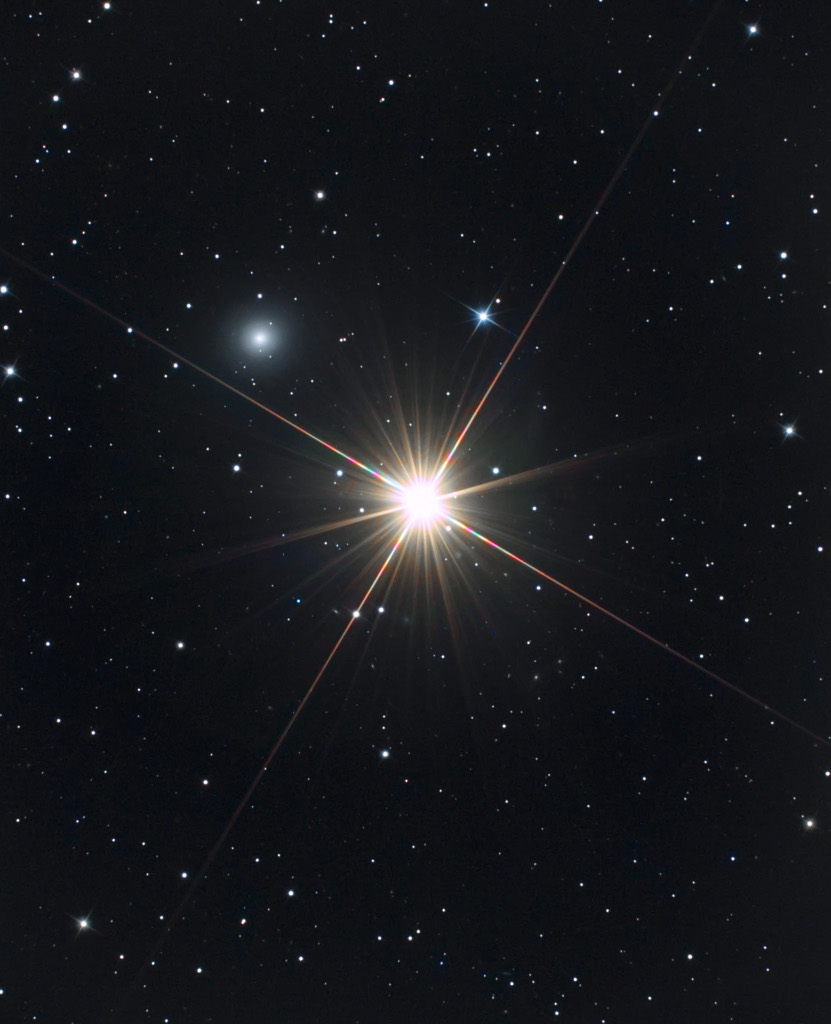 Mitten im Bild leuchtet ein heller Stern mit vielen Zacken, links darüber befindet sich ein verschwommener Fleck, der sich als Galaxie entpuppt.