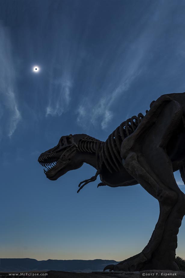 Hinter der Silhouette eines riesigen Dinosauriers und einigen Zirruswolken leuchtet die Korona am blauen Himmel, die Sonne selbst ist vom Mond verdeckt.