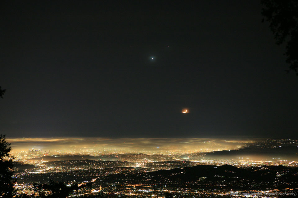 Über einer lichtverschmutzten Stadt, die bis zum Horizont reicht, leuchtet ein Sichelmond, links darüber stehen die Planeten Venus und Jupiter.