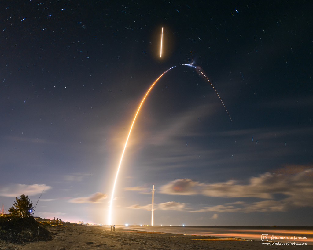 Am heiteren, leicht bewölkten Himmel zieht eine startende Rakete eine Leuchtspur, eine zweite Spur leuchtet am Scheitel des großen Bogens und eine dritte unten am Horizont.