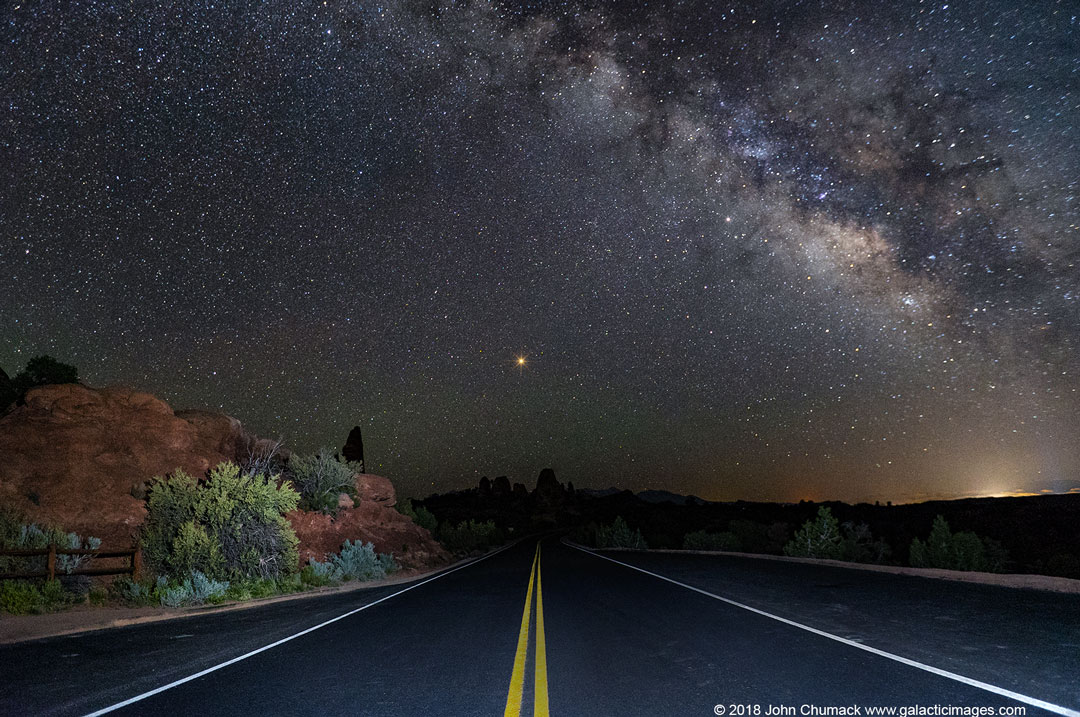 Die Straße führt scheinbar zum Planeten Mars. Links sind rötliche Felsen, die vom Scheinwerfer eines Autos beleuchtet werden, rechts oben stehen die Milchstraße und der Planet Saturn.