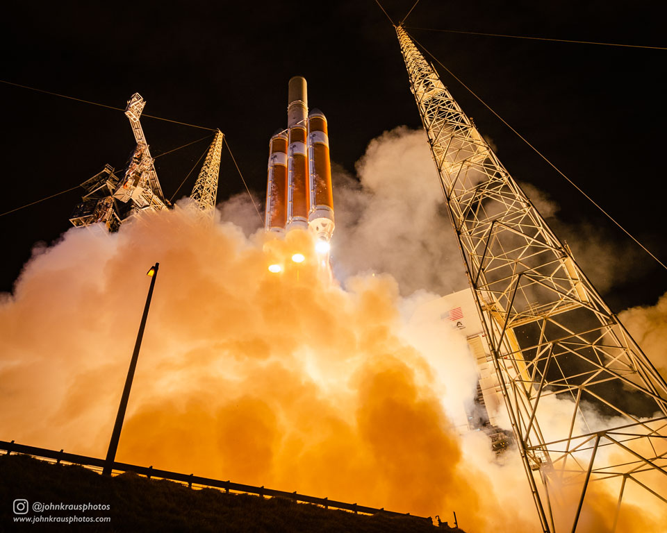 Über einer orangefarben leuchtenden Abgaswolke startet eine Trägerrakete mit der Raumsonde Parker Solar Probe in den Nachthimmel. Rechts ragt ein hoher Turm aus Stahlrohren auf.