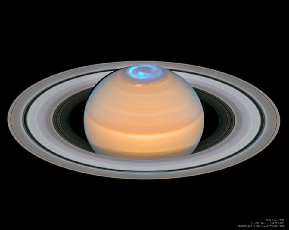 Wir sehen den Planeten Saturn schräg von oben, die Ringe bilden ein breites Oval, das oben und unten über den Planeten hinausreicht. Oben am Pol leuchtet ein cyanblaues Polarlicht in Form einer Spirale.