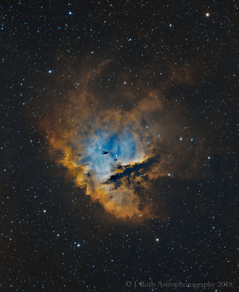 Der Pac-Man-Nebel im Bild leuchtet hier innen blau und ist von einem Rand aus orange leuchtenden Nebeln umgeben.