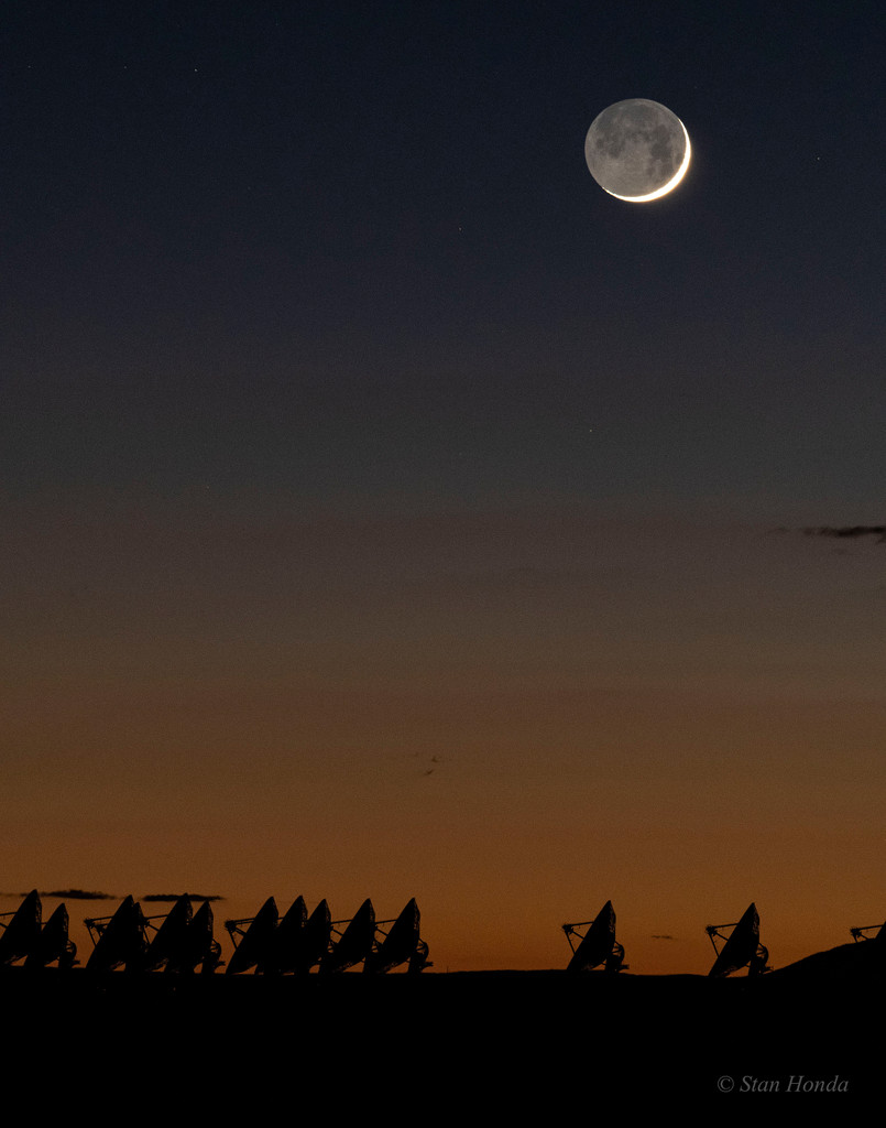 Am Horizont ragen die Silhouetten zahlreicher Radioteleskope auf. Der Himmel dahinter leuchtet dunkelrot und wird nach oben hin dunkelblau. Rechts oben leuchtet ein junger Sichelmond, dessen Nachtseite von der Erde beleuchtet wird.