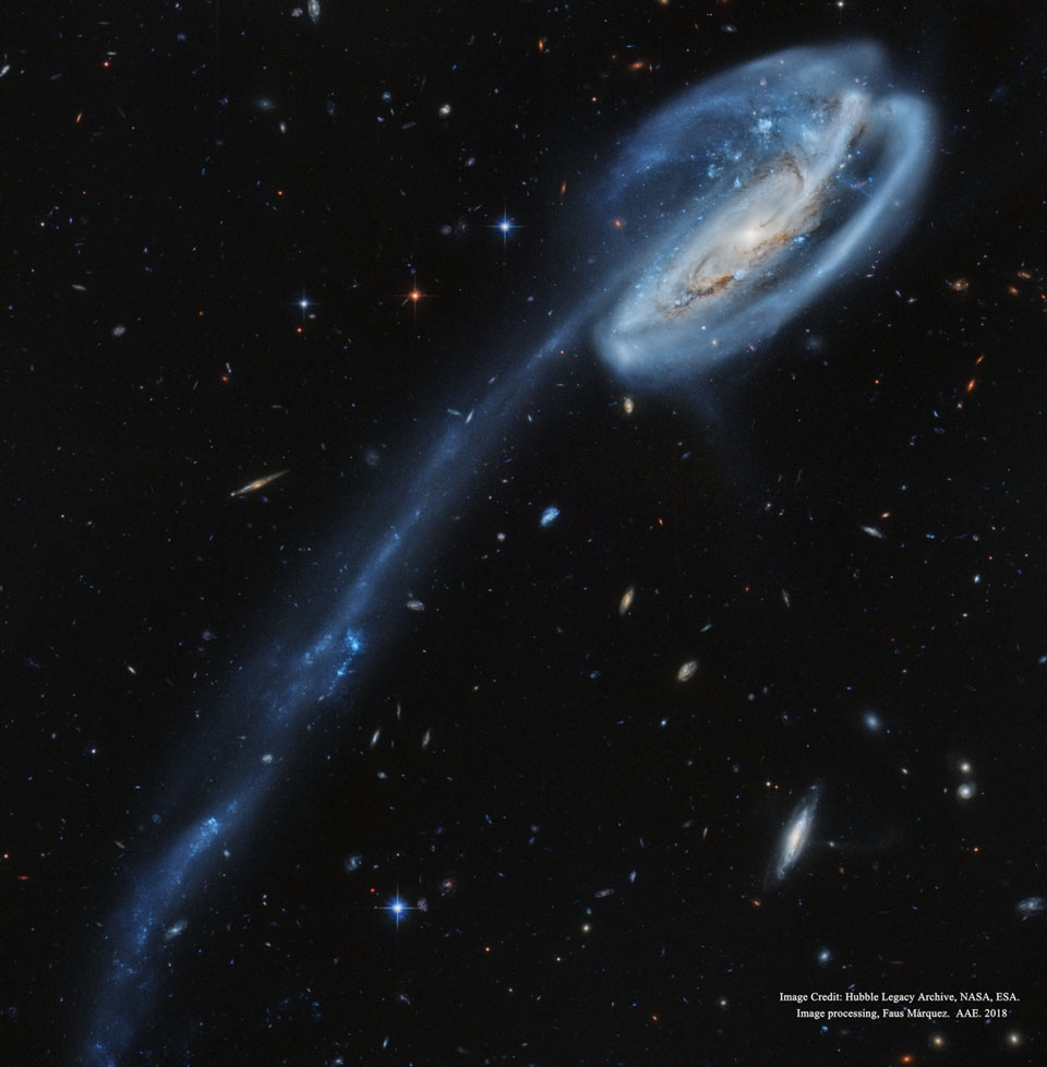 Rechts oben ist eine Galaxie, die einen sehr dreidimensionalen Eindruck macht. Die Spiralarme erscheinen in zwei Ebenen gewickelt, nach rechts unten verläuft ein Strang aus Sternen und blauen Sternhaufen. Im Hintergrund sind zahlreiche Galaxien verteilt.