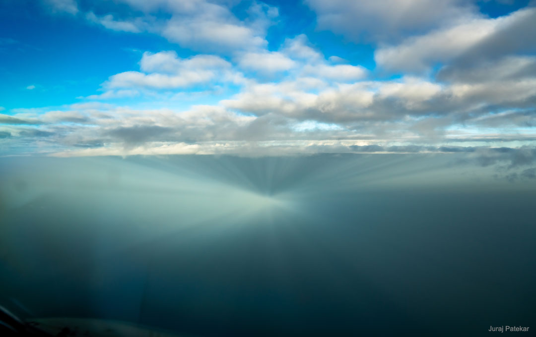 Beim Blick aus dem Fenster eines Flugzeugs waren diese Gegendämmerungsstrahlen zu sehen. Es sind Strahlenbüschel, die gegenüber der Sonne wieder zusammenlaufen. Oben ist der Himmel wolkig und blau, unten ist grauer Nebel.