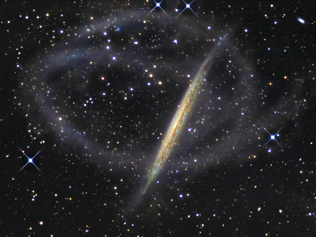 Hinter einer on der Seite sichtbaren Galaxie, die leicht verkrümmt ist, verlaufen schlierenförmige Sternströme.