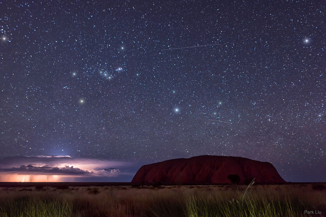 Hinter Uluru, einem Weltnaturerbe in Zentralaustralien, tobt ein Gewitter, am Himmel leuchtet das Sternbild Orion.