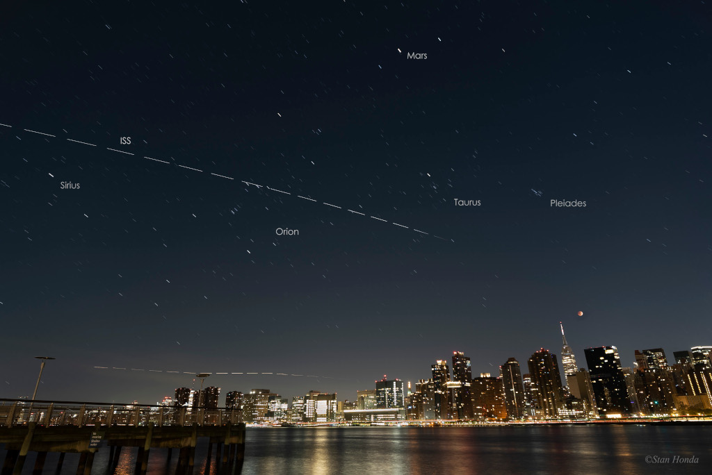 Das Bild zeigt eine Mondfinsternis über Manhattan, am Himmel sieht man die Plejaden, das Sternbild Orion und die Leuchtspur der Internationalen Raumstation.