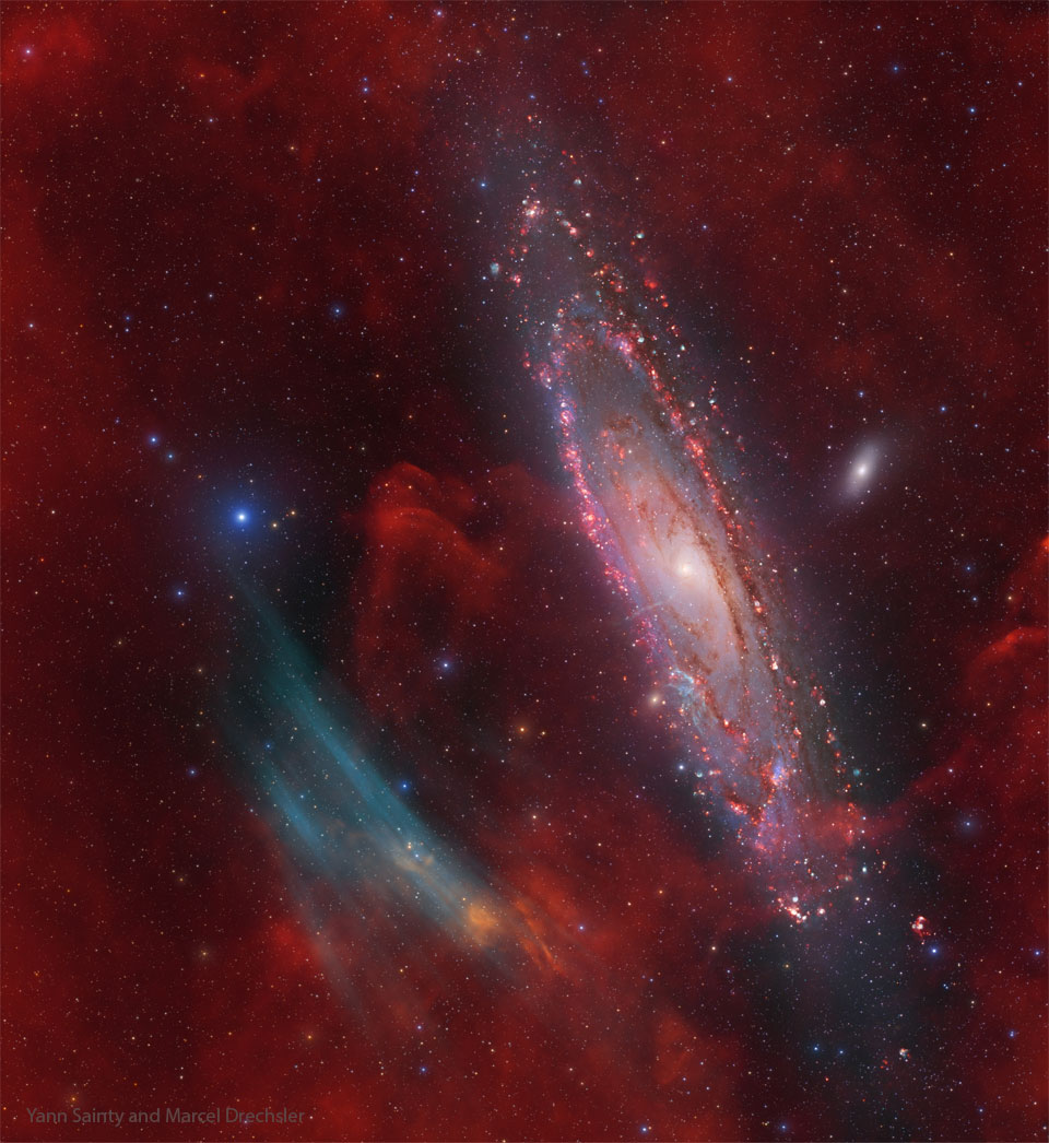Das Amateurbild zeigt die Andromedagalaxie umgeben von rotem Leuchten von Wasserstoff. Links neben der Galaxie sind blau leuchtende Bögen aus Sauerstoff. Beschreibung im Text.