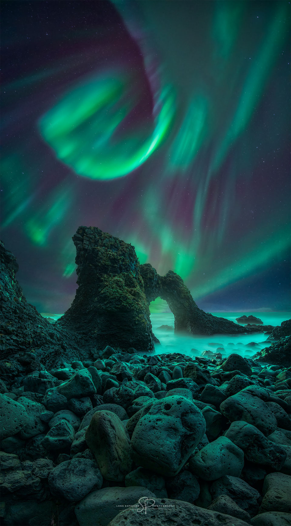 Über einer fantastischen Küste mit Steinen und einem Felsbogen wogt ein gewundenes lebhaftes Polarlicht.