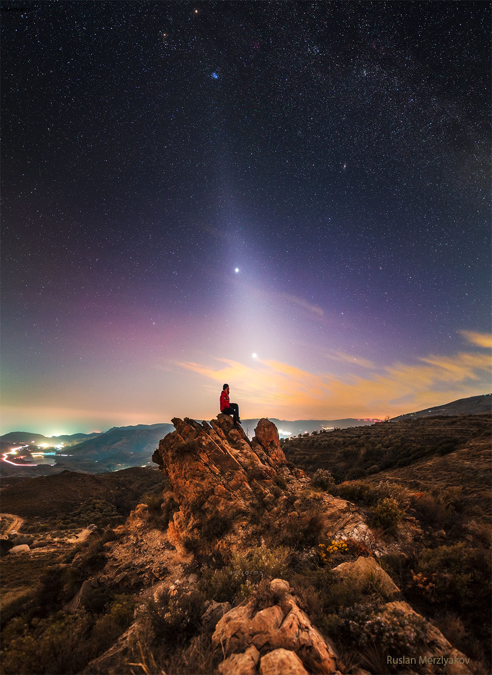 MAuf einem Felsen sitzt eine Person unter einem ungewöhnlichen Himmel. Am Himmel leuchtet ein helles, diffuses Band, das sich bis zum Horizont erstreckt und durch zwei helle Punkte, Jupiter und Venus, verläuft. Die Plejaden Sternhaufen ist über ihnen zu sehen.