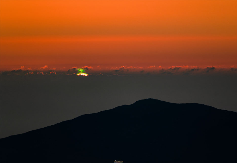 Hinter der Silhouette eines Berges ist eine Wolkendecke zu sehen, die unten grau ist und oben einen geraden, gekräuselten Rand hat, hinter dem die Sonne versinkt und dabei oben einen grünen Blitz bildet. Über der Wolkendecke ist der Himmel orangefarben.