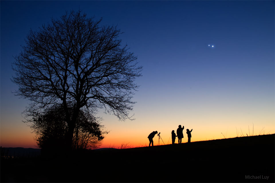 Am blauen Abendhimmel leuchten nach Sonnenuntergang zwei helle Planeten, über dem dunklen Horizont leuchtet Abendrot, davor sind die Silhouetten eines Baumes zu sehen sowie vier Menschen.