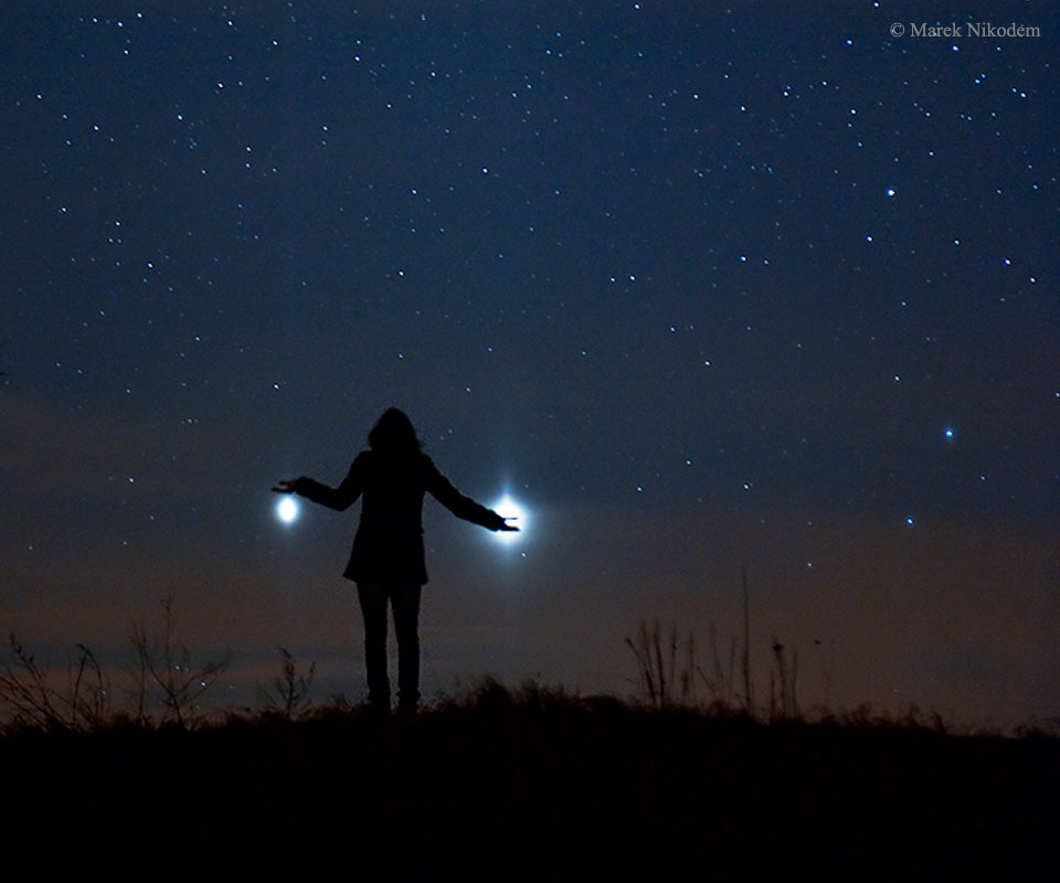Eine junge Frau hält scheinbar die Planeten Venus und Jupiter, die am sternklaren Himmel leuchten.