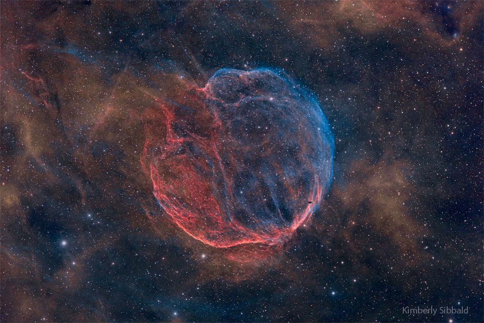 In der Mitte des Bildes mit Sternennebeln leuchtet ein runder Nebel aus Fasern, der auf einer Seite blau und links unten rot leuchtet.