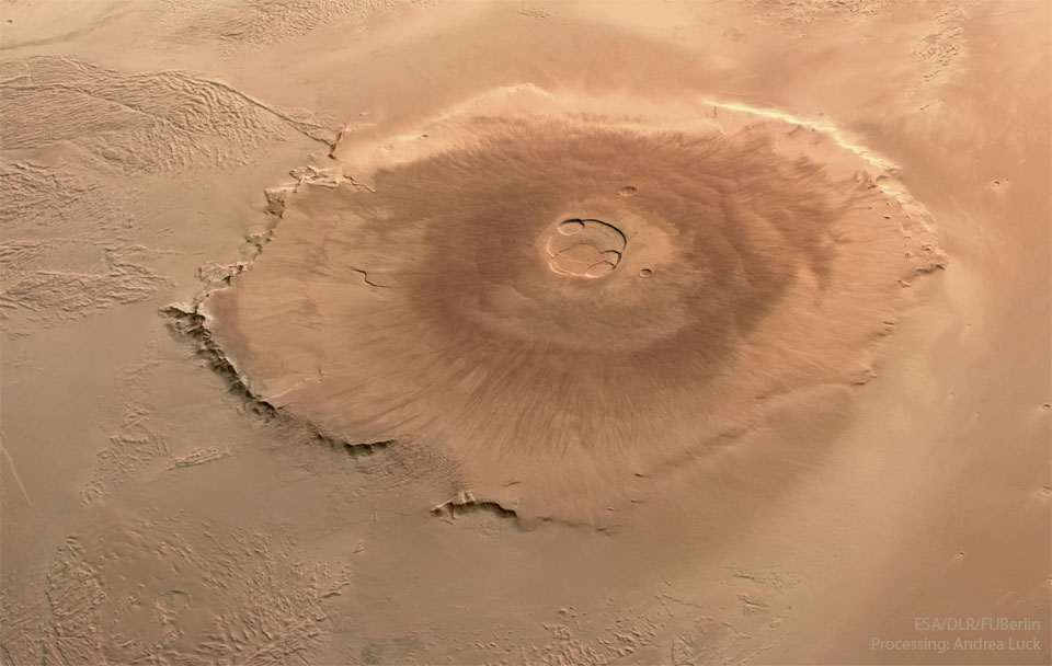 In der Bildmitte liegt der Schildvulkan Olympus Mons, er ist der größte Vulkan im Sonnensystem. Die Marsoberfläche ist rötlichbraun, der Vulkan wirkt wie eine flache Flade mit einer goßen Vertiefung in der Mitte. Rund um den Krater am Gipfel befindet sich eine dunklere Region.