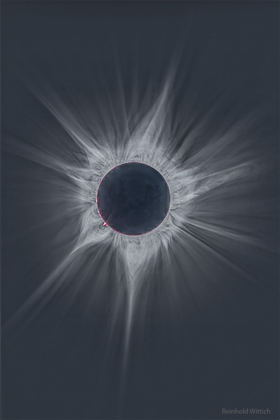 Der dunkle Neumond bedeckt die Sonne und ist von hellen Strahlen der Sonnenkorona umgeben. Über den Rand ragen zarte rosarote Protuberanzen.