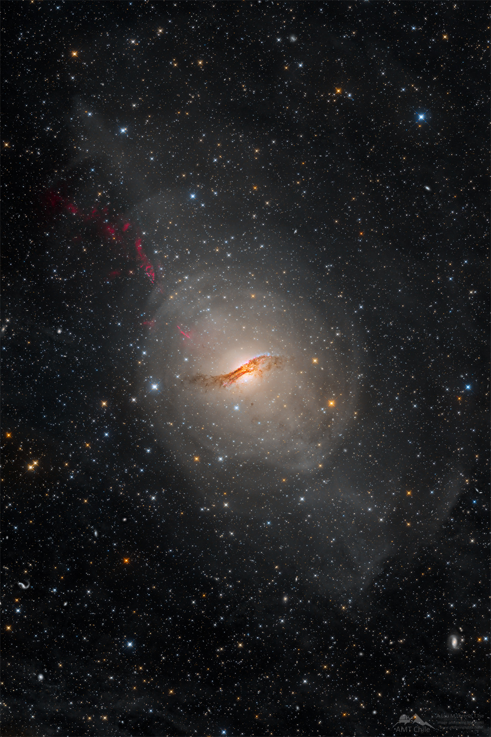 Die Galaxie Zentaurus A leuchtet in der Mitte dieses Sternenfeldes, davor verläuft ein dunkles Staubband. Nach links oben strömt ein roter Strahl.