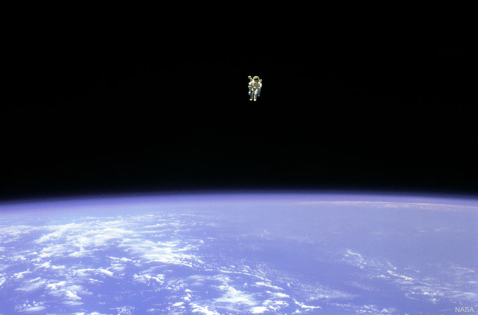 Über der blauen Erde mit wenigen Wolken, die oben gekrümmt erscheint, schwebt im schwarzen All in großer Entfernung ein Astronaut ohne Leine.