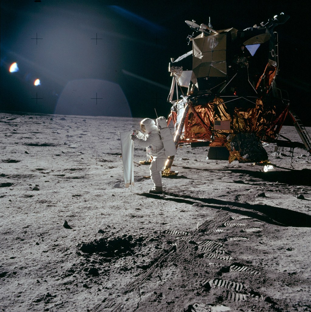 Das Bild von der Mondoberfläche zeigt links das Mondlandemodul, in der Mitte stellt der Astronaut Buzz Aldrin ein Sonnenwind-Experiment auf. Der Himmel ist schwarz, der Boden von dunklem grauem Staub bedeckt.