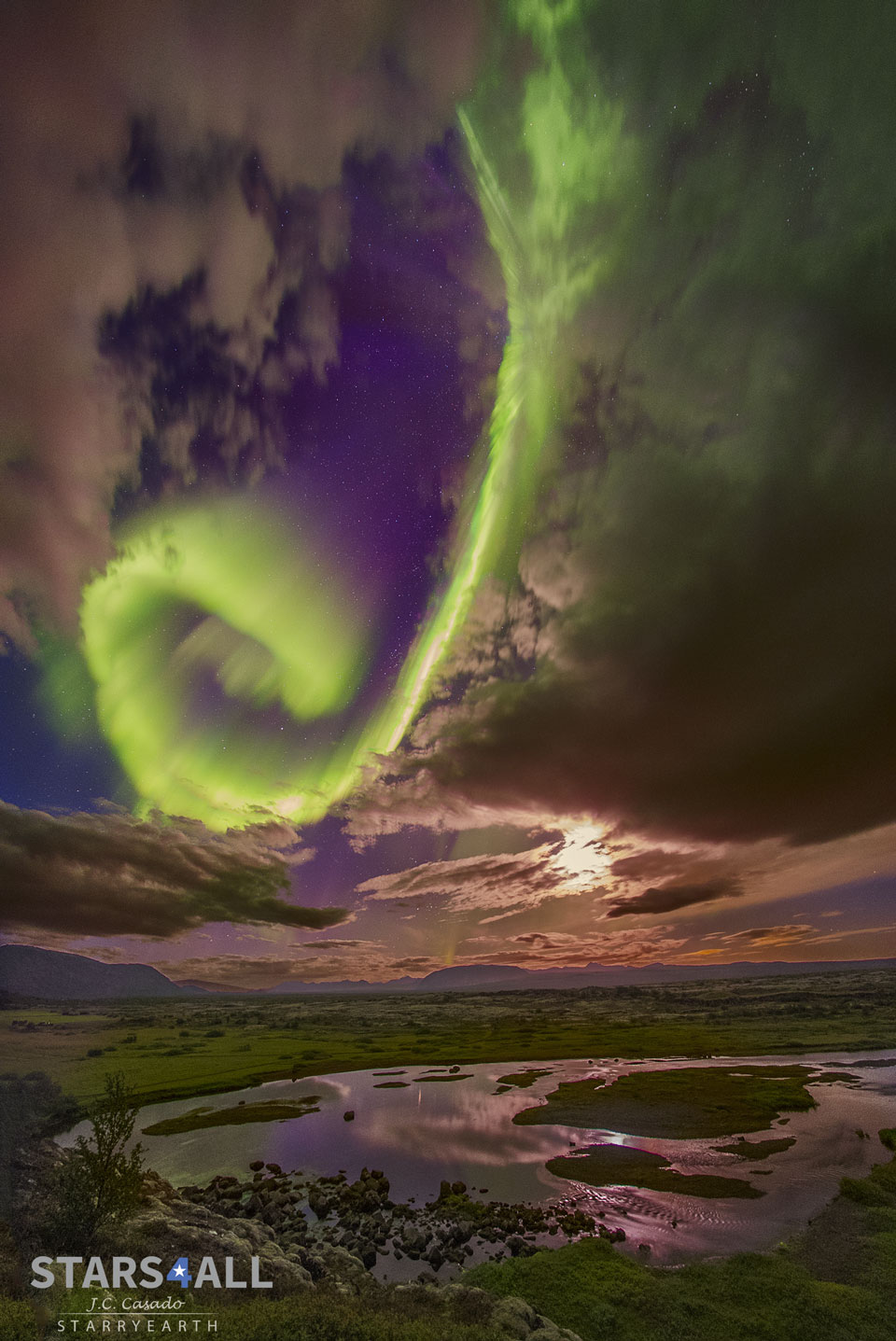 Über einer duklen, düsteren Landschaft mit wilden Wolken wölbt sich ein grünes Polarlicht in Form einer Spirale.