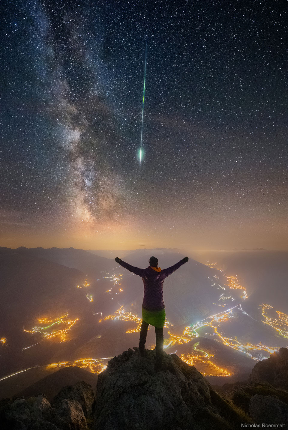 Im Vordergrund jubelt ein Himmelsbeobachter über einen hellen Meteor, der neben der Milchstraße leuchtet. Im Tal sieht man die Lichter von Siedlungen.