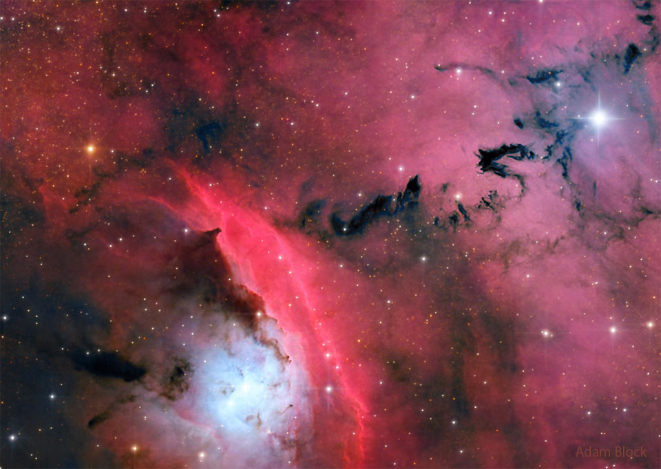 Das rötlich beleuchtete Bild wirkt chaotisch, quer im Bild schlängeln sich dunkle Wolkenbänder, links unten leuchtet ein heller blauer Neel um einen Stern, daneben ist ein rot leuchtender Nebelstreifen.