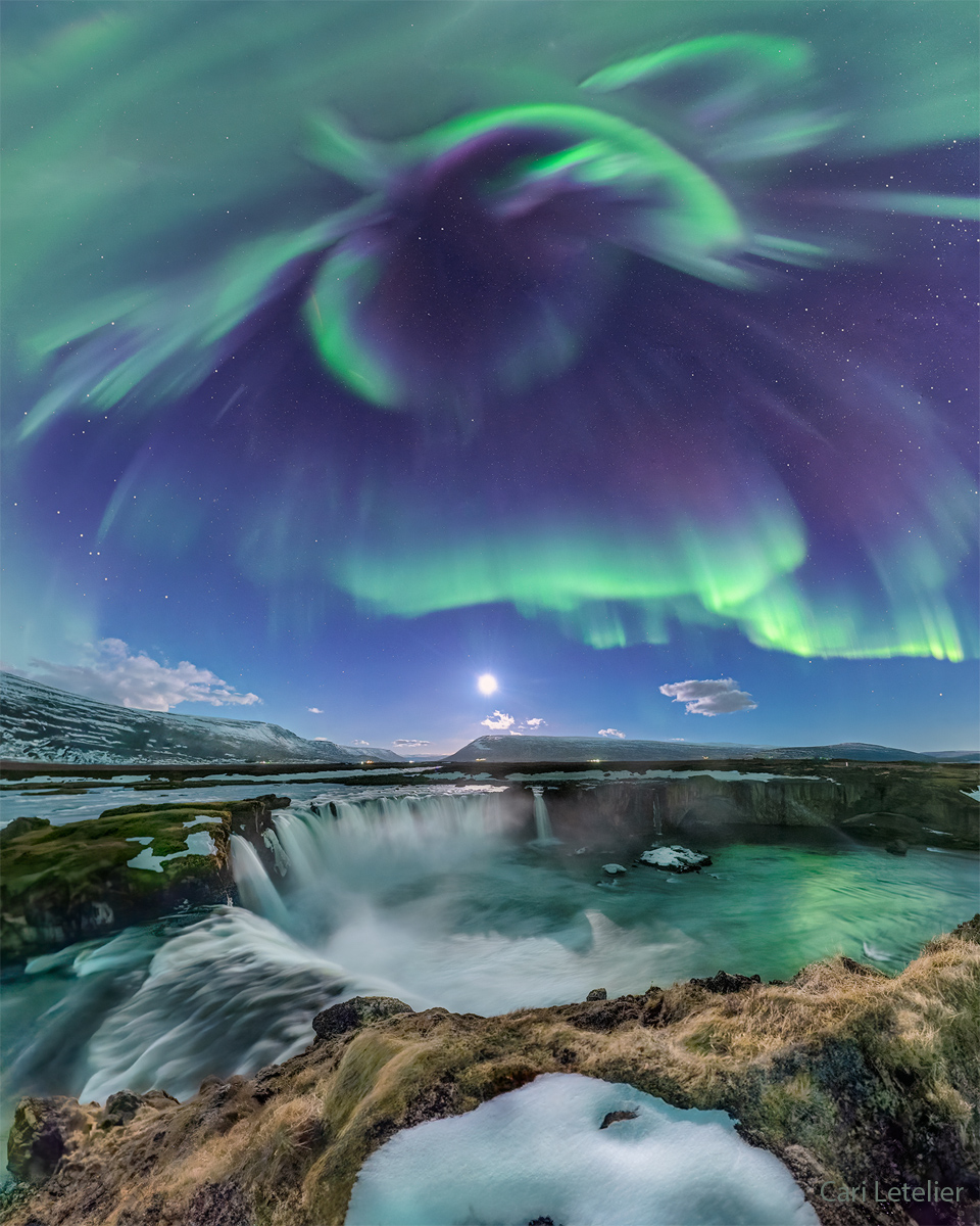 Über einem Wasserfall leuchtet ein grünes Polarlicht,im Hintergrund steht über dem Horizont der Mond.