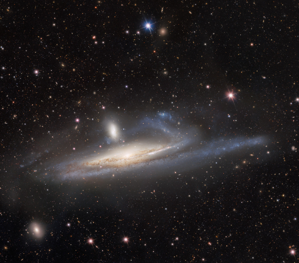 Von einer seitlich sichtbaren Spiralgalaxie steigt ein Arm aus Sternen auf, darüber schwebt eine kleine, senkrechte Galaxie. Im Bild sind weitere Galaxien und Sterne zu sehen.