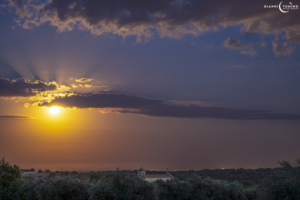 Über einer Waldlandschaft verläuft am sonst klaren Himmel eine waagrechte Wolke, unter der ein helles Licht hervor leuchtet. Unter der Wolke wirkt der Himmel orangefarben, über der Wolke ist er dunkelblau, und über dem Licht - dem Mond - sind Strahlenbüschel.