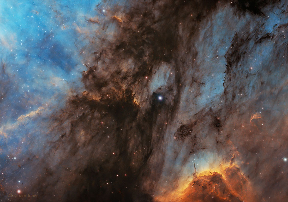Das Bild ist von einem bläulich leuchtenden Nebel gefüllt, über den in der Mitte ein dunkler Staubwolkenstreifen verläuft. Rechts unten liegen zwei orangebraune Nebelhäufchen.